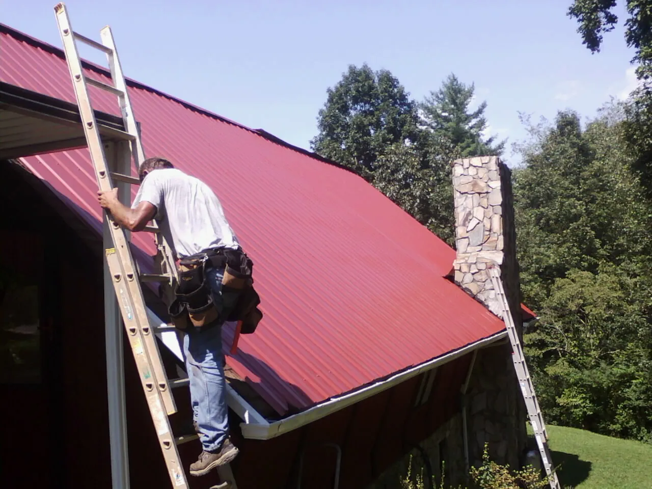 Roofer on a ladder beside a red metal roof and stone chimney — Soffit & Fascia Repair by Roofing Brothers in Hidden Valley